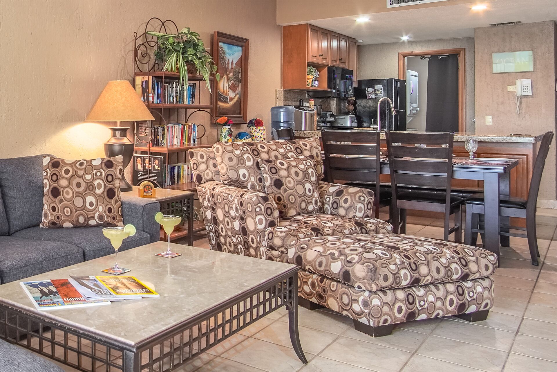 Ein Wohnzimmer mit Sofa, Hocker und Couchtisch, ein Essbereich mit Stühlen, eine Küche mit Schränken und Geräten sowie ein Bücherregal mit Büchern und einer Pflanze.