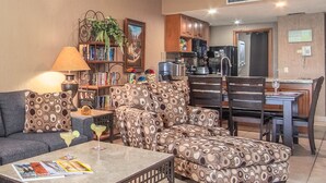 A living room with a sofa, ottoman, and coffee table, a dining area with chairs, a kitchen with cabinets and appliances, and a bookshelf with books and a plant. - Princesa Resort D-307 Pelican's Post Ocean View Chic Condo (Puerto Peñasco)