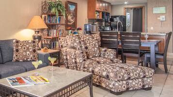 Ein Wohnzimmer mit Sofa, Hocker und Couchtisch, ein Essbereich mit Stühlen, eine Küche mit Schränken und Geräten sowie ein Bücherregal mit Büchern und einer Pflanze.