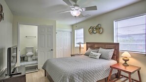 Un dormitorio con cama, ventilador de techo, mesita de noche con lámpara, cómoda, televisor y un baño visible a través de una puerta abierta.