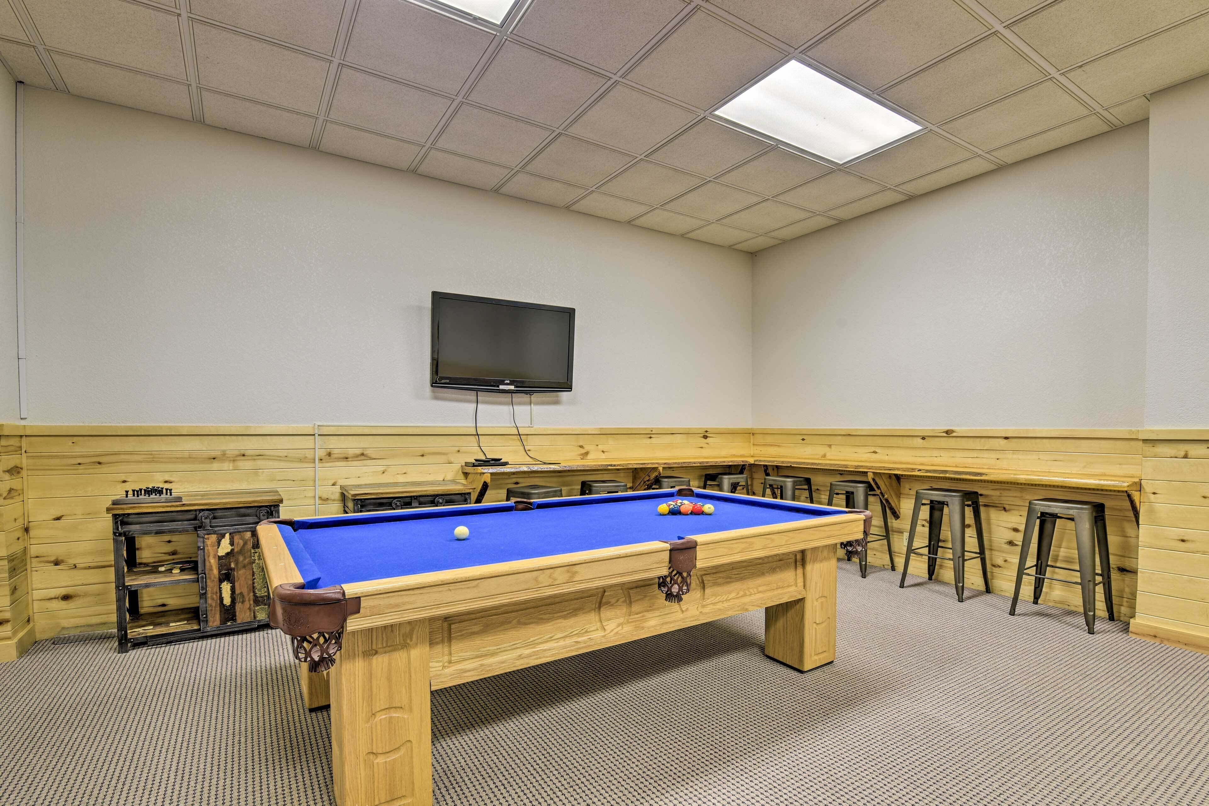 A pool table with a blue felt surface, a television mounted on the wall, and wooden bar stools along the wall.
