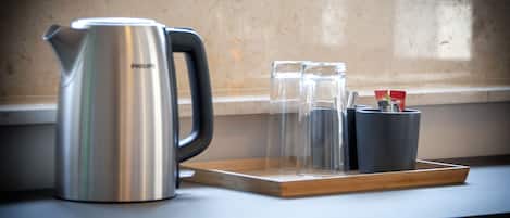 Une bouilloire en acier inoxydable, posée sur un plateau en bois, avec deux verres et une tasse, le tout sur un comptoir près d’une fenêtre.