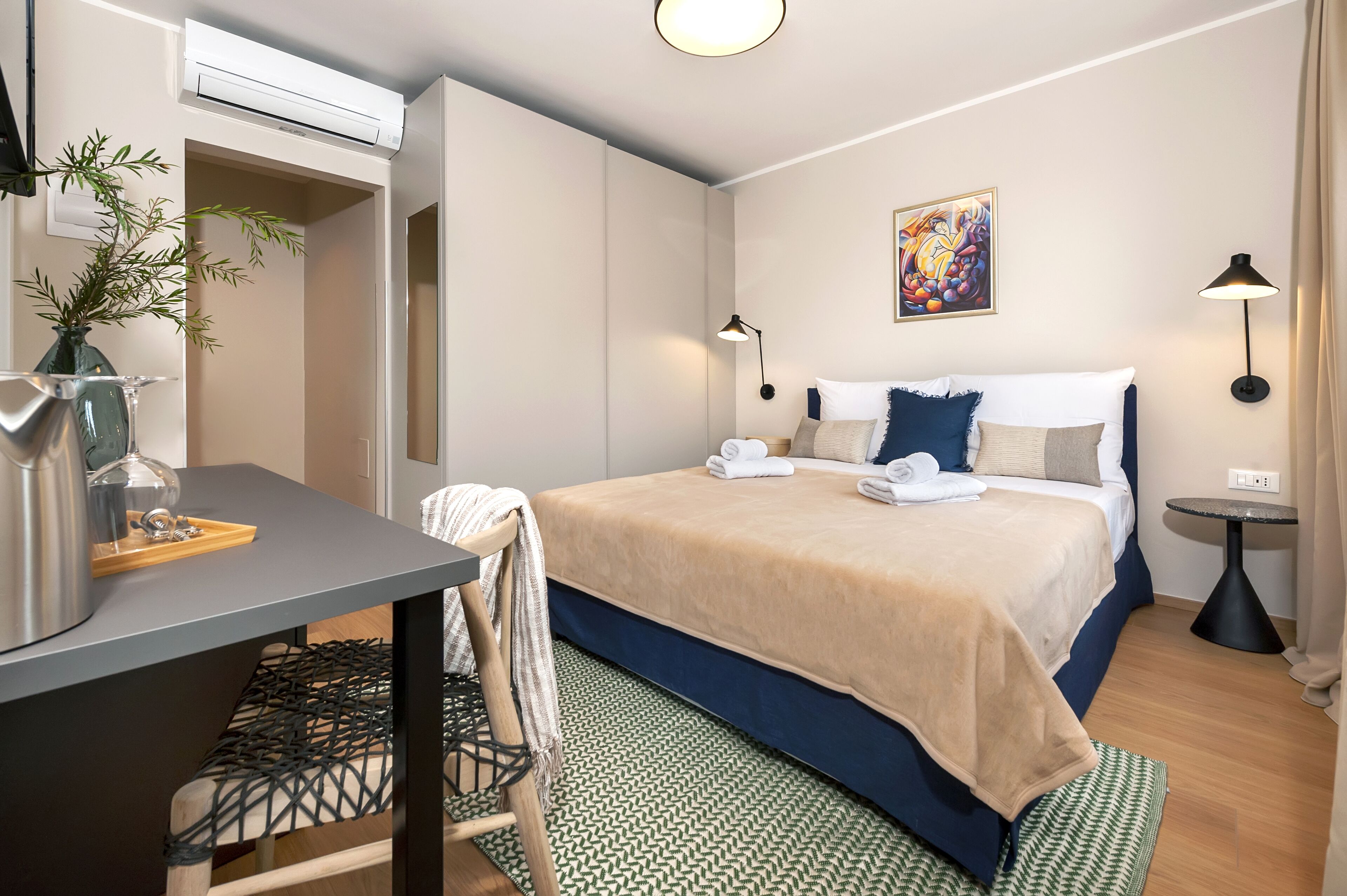 Une chambre d’hôtel avec un lit, un bureau, une chaise, une petite table, une lampe et un tableau au mur.