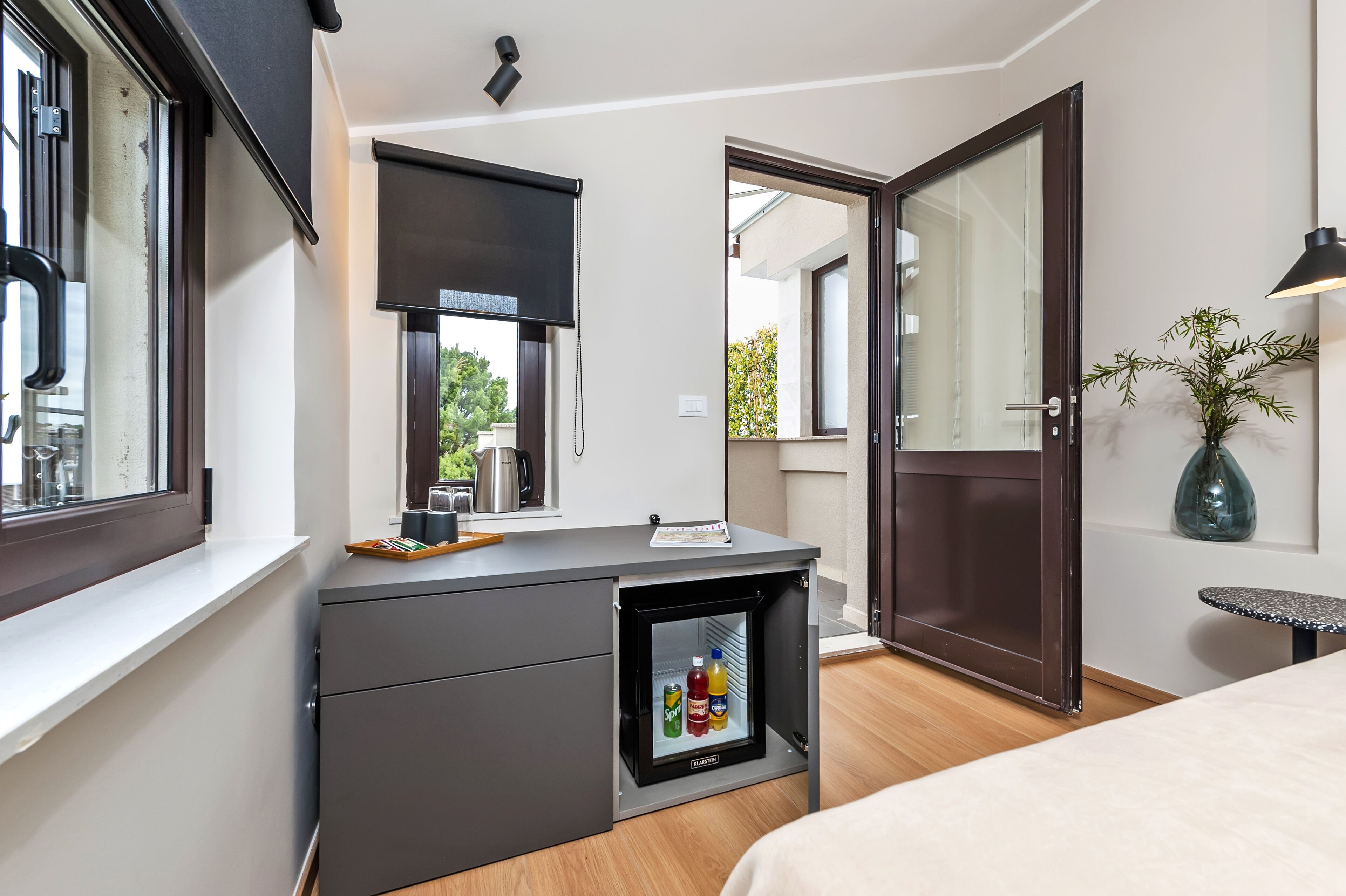 A compact room with a built-in mini-fridge, a desk with a computer, a bed, and a door leading to an outdoor area.