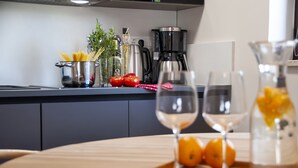 Una encimera de cocina con una cafetera, una olla de espagueti y tomates frescos. Hay dos copas de vino sobre una bandeja con naranjas.