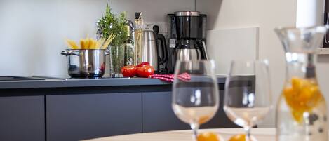 A kitchen counter with a coffee maker, a pot of spaghetti, and fresh tomatoes. There are two wine glasses on a tray with oranges.