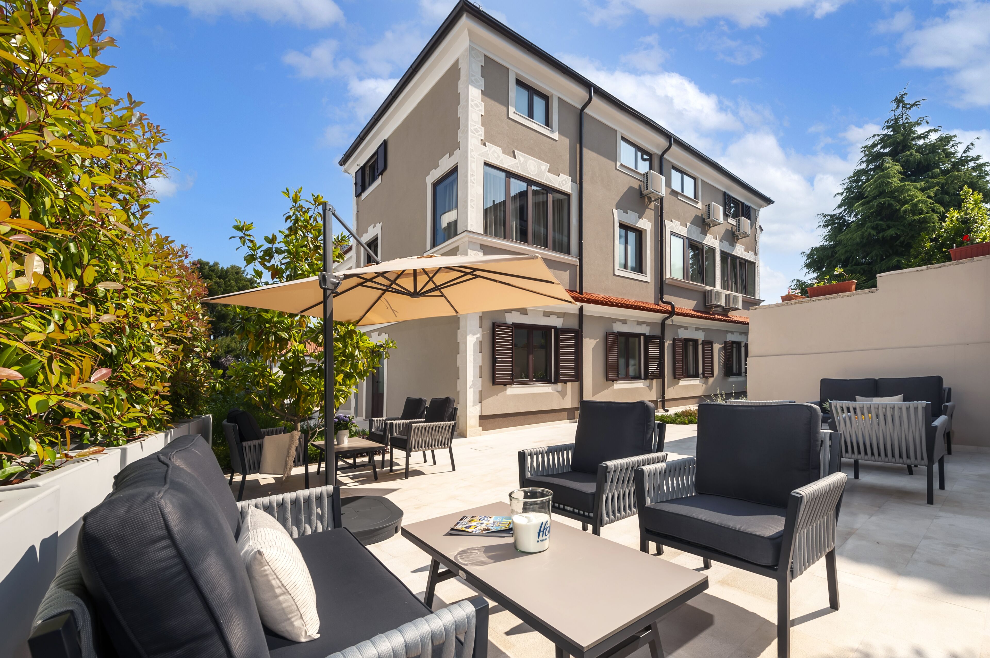 Un patio al aire libre con diferentes espacios de descanso, incluyendo sofás y sillones, y un gran paraguas que ofrece sombra.