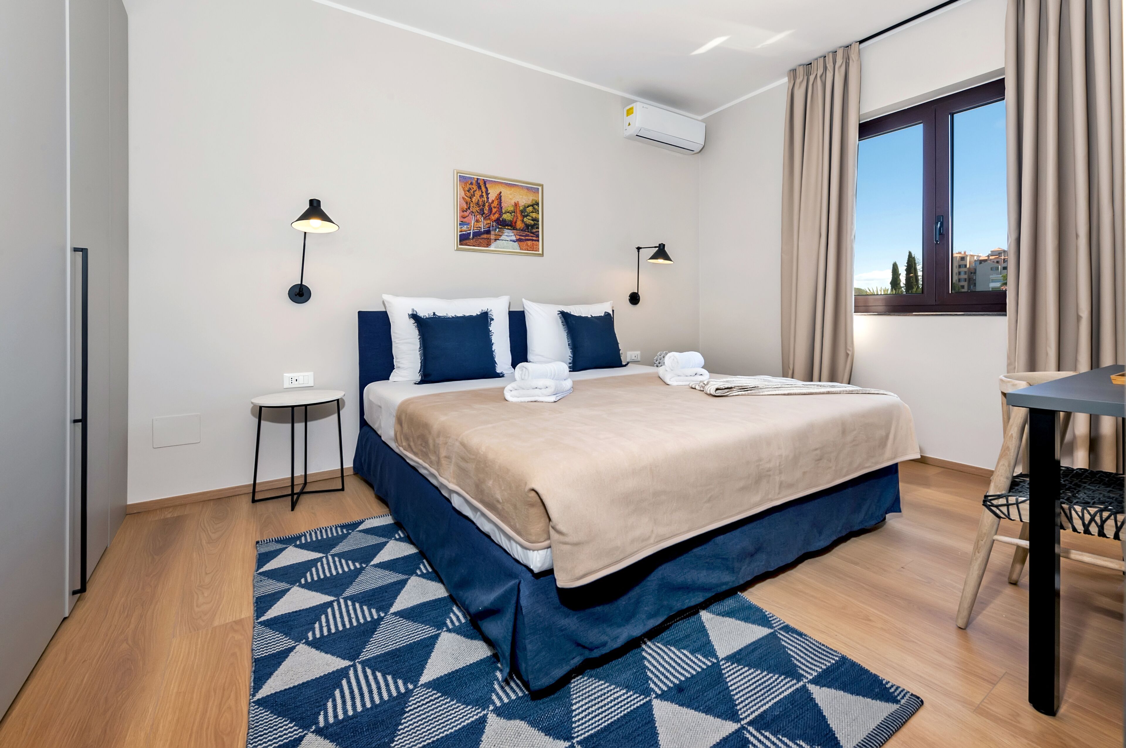 A modern hotel room with a large bed, a small table, a chair, a wall-mounted air conditioner, and a window with curtains.