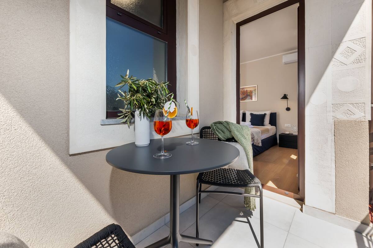 A balcony with a round table, two chairs, a potted plant, and two glasses of red wine.