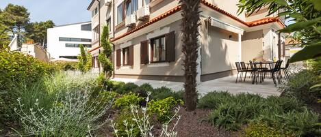 Un edificio de dos pisos con un balcón, rodeado de abundante vegetación y una palmera.