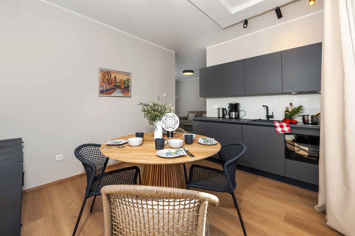 A modern kitchen with dark cabinets, a dining area with a round table, and a wicker chair.