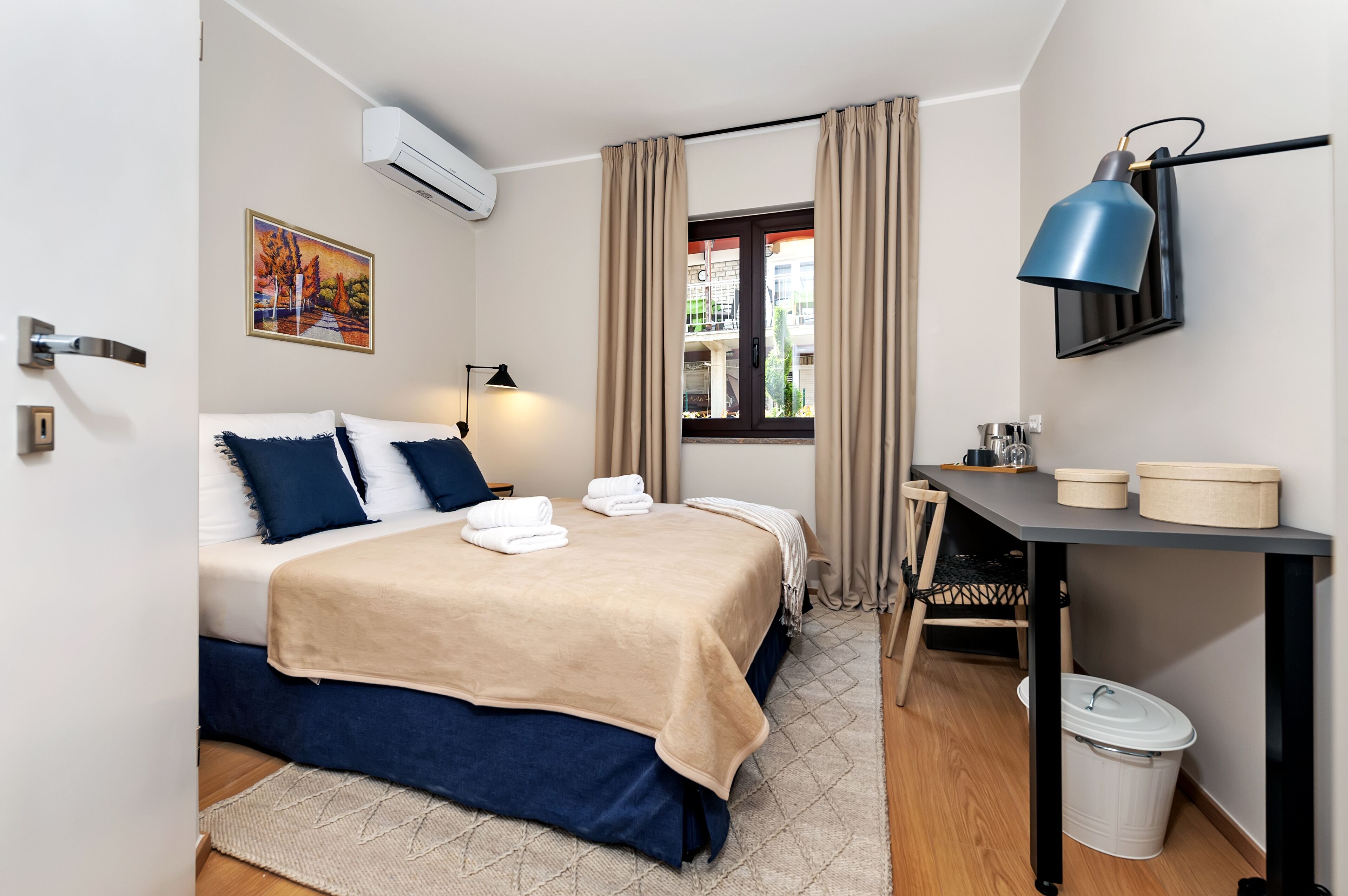 Een slaapkamer met een bed, een bureau, een stoel, een raam en een prullenbak.