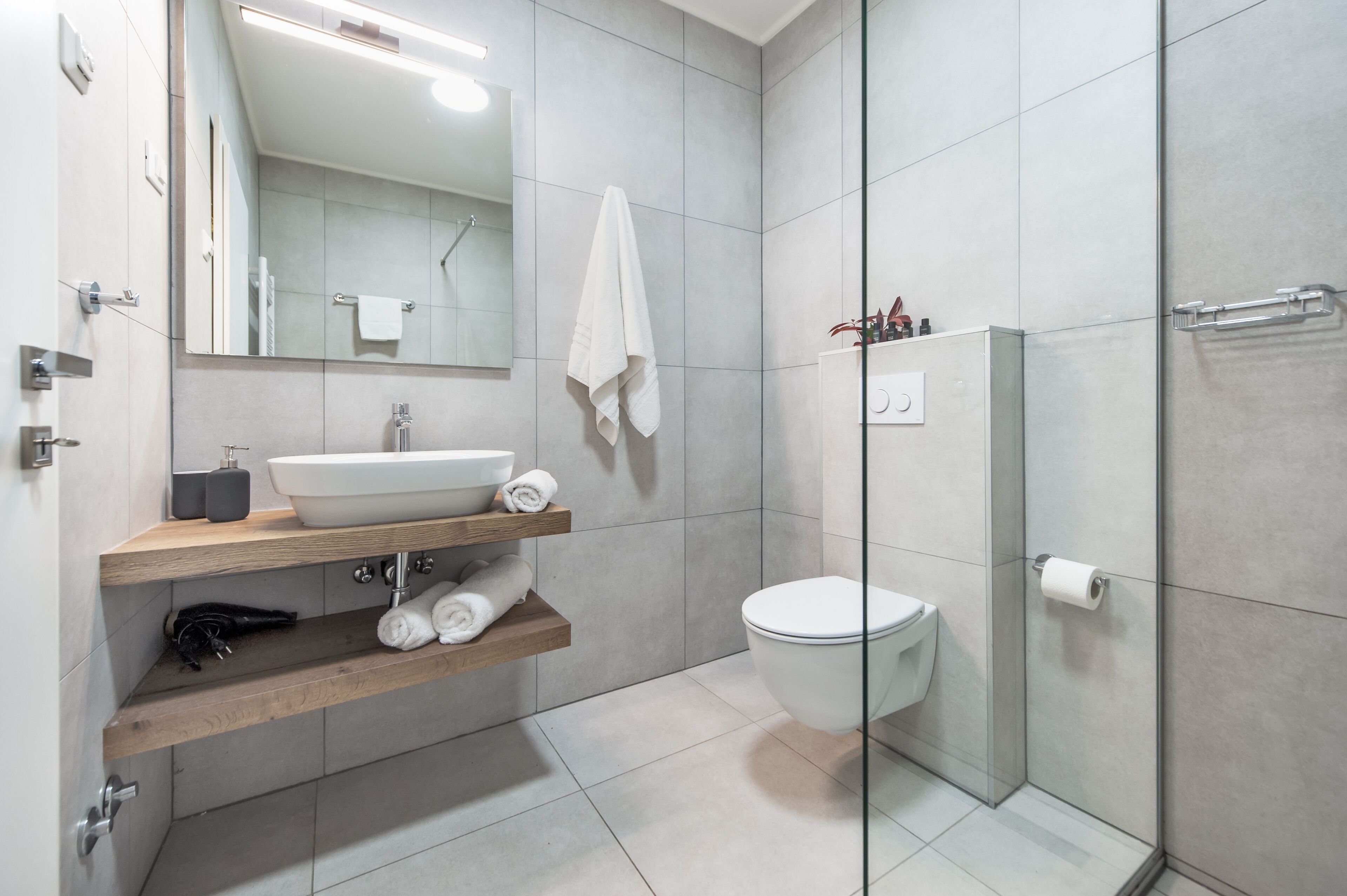 Ein modernes Badezimmer mit einem weißen Waschbecken, einem Spiegel und einer Toilette. Holzregale mit Handtüchern und einer Duschkabine aus Glas.