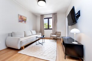 A modern living room with a sofa, a small table, a chair, a TV mounted on the wall, and a window with curtains.