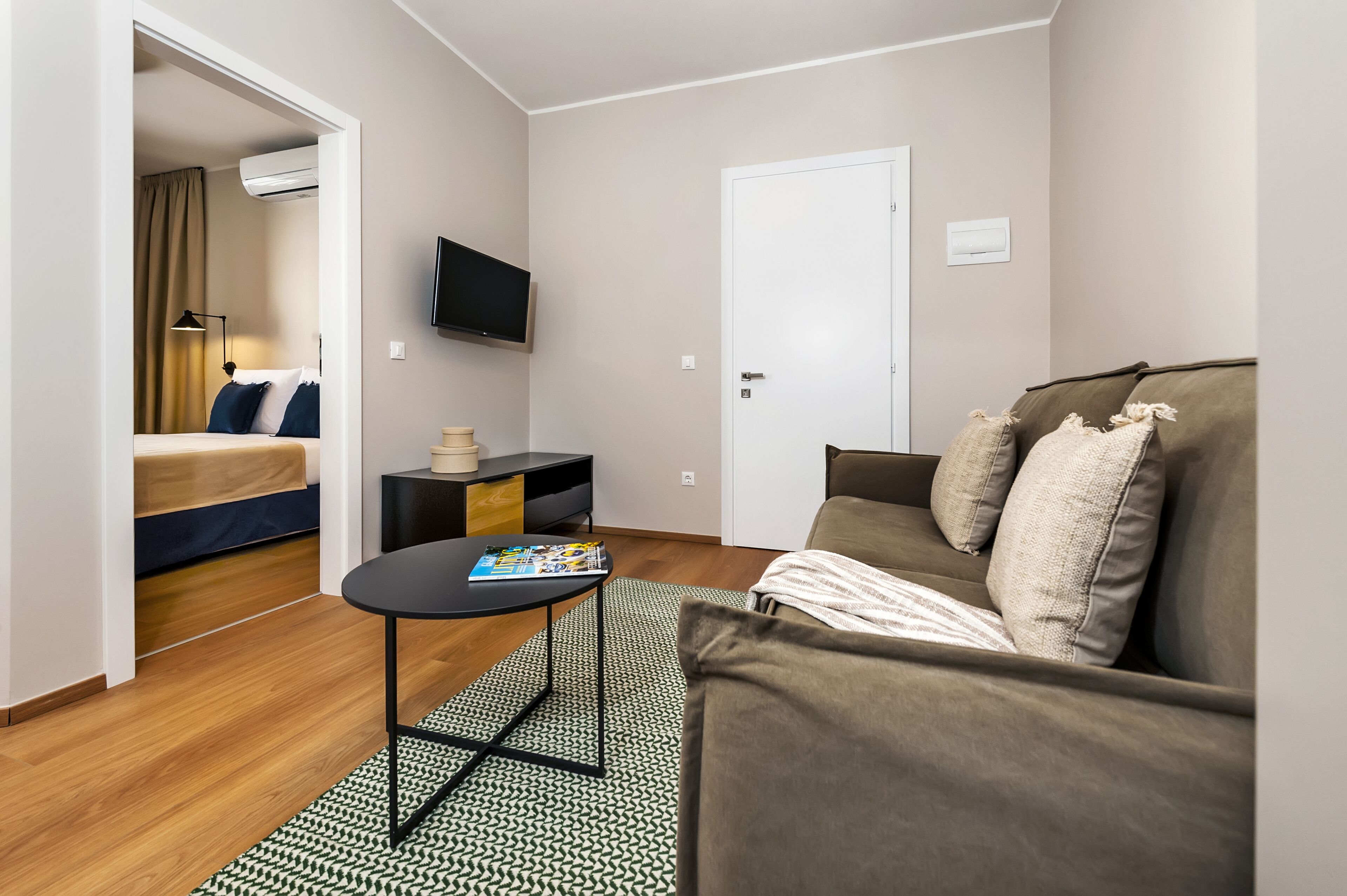 A compact hotel room with a bed, a sofa, a small table, a TV, and a door.