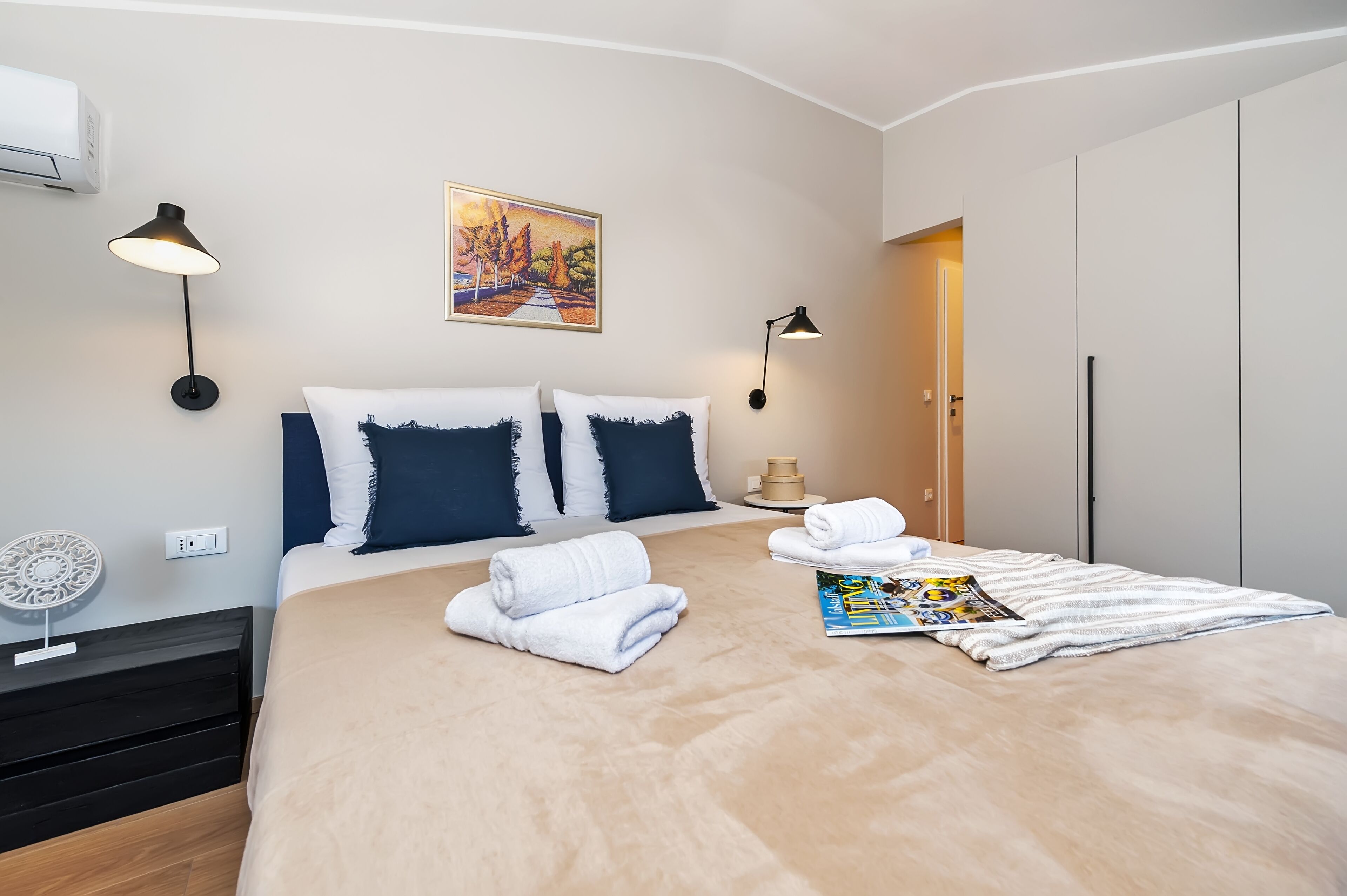 Una cama bien hecha con almohadas blancas y azul marino, una revista y toallas enrolladas. Hay dos lámparas de pared y un cuadro sobre la cama.