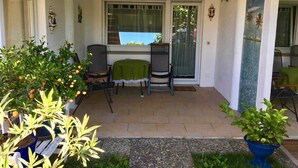 Eine überdachte Terrasse mit zwei Stühlen und einem Tisch, ein Fenster mit Blick und Kübelpflanzen.