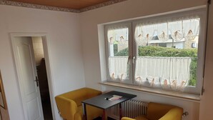 Ein Zimmer mit rotem Teppich, zwei gelben Stühlen, einem kleinen Tisch, einem Heizkörper und einem Fenster mit Spitzenvorhängen.