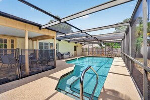 Une piscine extérieure couverte, protégée par une structure en verre, un espace patio avec des chaises, et une vue sur une clôture en bois et des arbres.