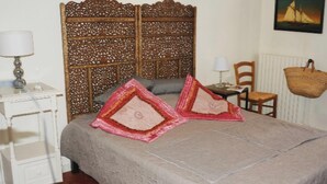 Een slaapkamer met een bed, twee kussens, een nachtkastje, een lamp, een stoel, een raam en een wandmand.