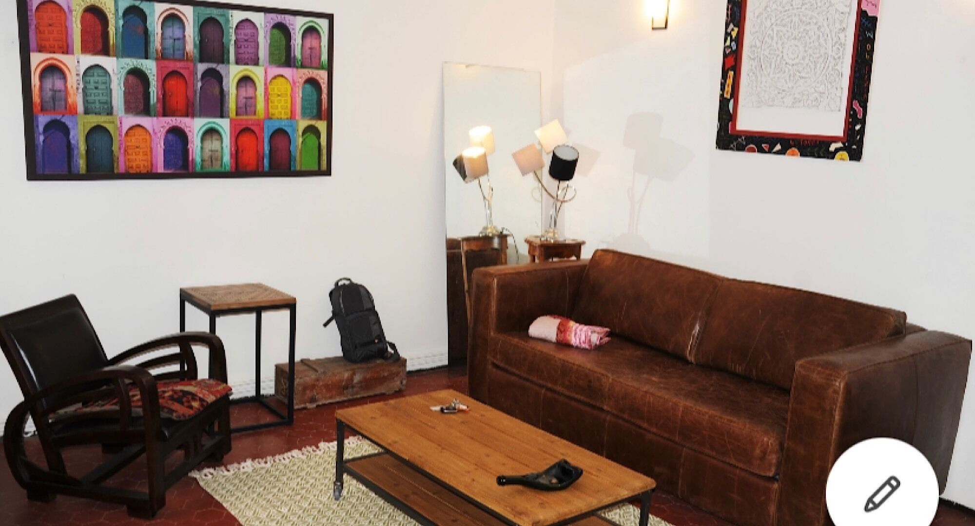 Un salon avec un canapé marron, une table basse en bois, une petite table, une chaise, un sac à dos et un tableau coloré représentant des portes.