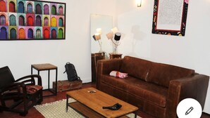 Een woonkamer met een bruine bank, een houten salontafel, een klein tafeltje, een stoel, een rugzak en een kleurrijk schilderij van deuren.