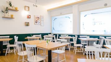 Un comedor con sillas y mesas blancas, estanterías de madera con objetos decorativos, y un amplio ventanal con vistas.