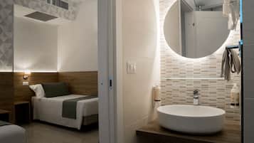 A modern hotel room with a bed, bedside table, and a bathroom with a round mirror, sink, and towel rack.