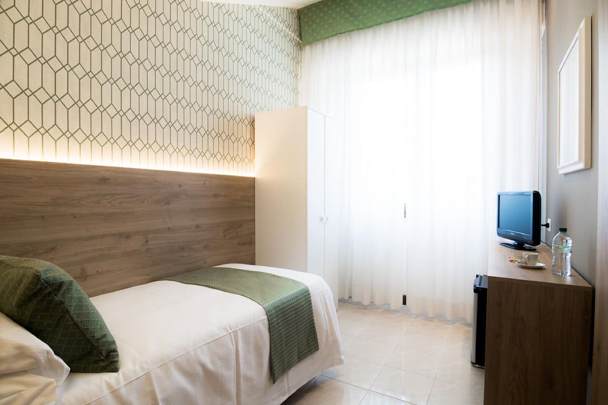 Hotellihuone, jossa on puinen sängynpääty, vihreillä tekstiileillä varustettu sänky, yöpöytä, jossa on televisio ja vesipullo, sekä ikkuna, jossa on ohuet verhot.