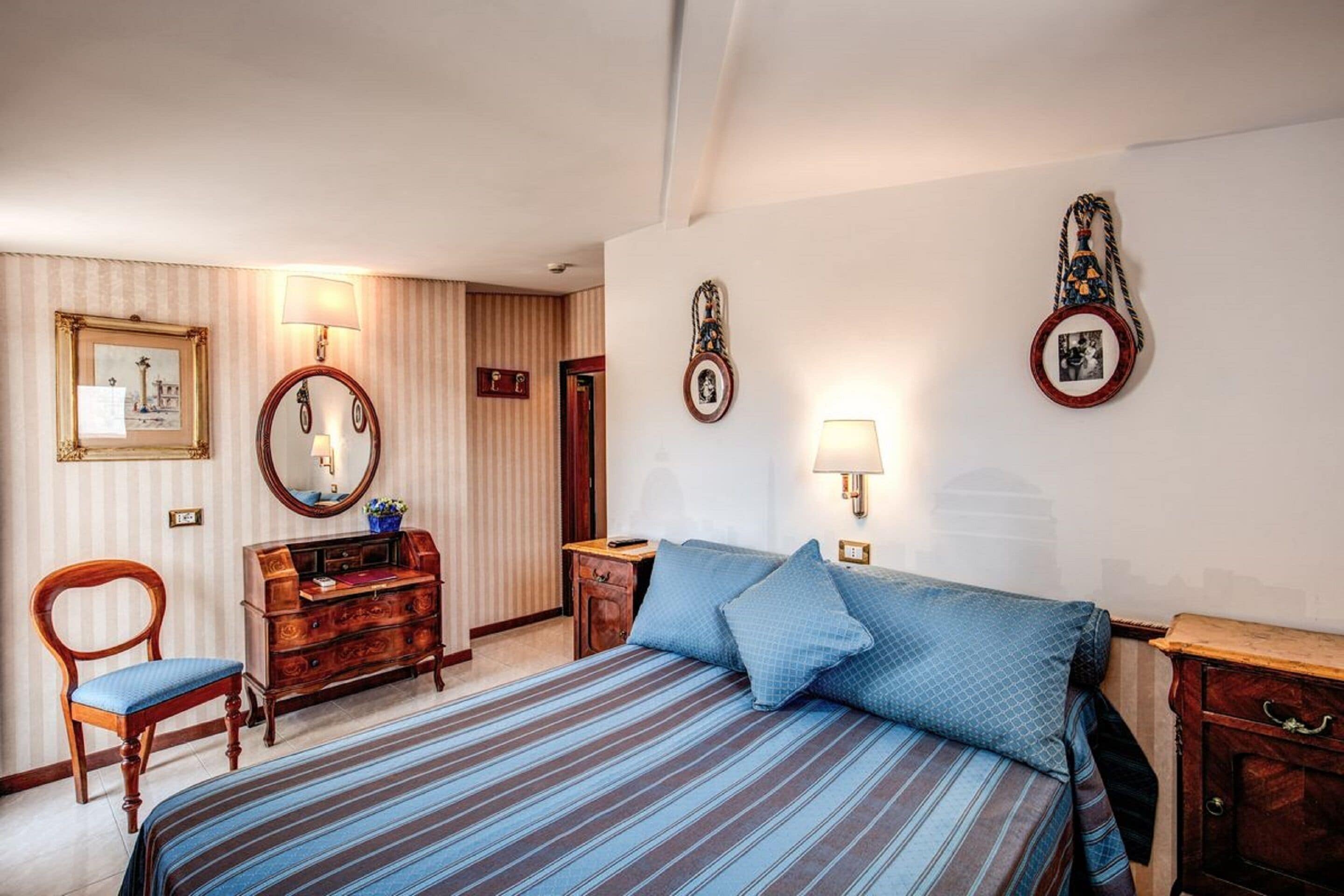 A hotel room with a bed, a wooden dresser, a chair, a desk, and wall-mounted lamps.