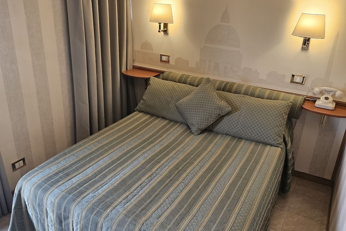 Hotellihuone, jossa on sänky, kaksi yöpöytävalaisinta, ilmastointilaite ja seinällä kaupunkimaisemaa esittävä seinämaalaus.