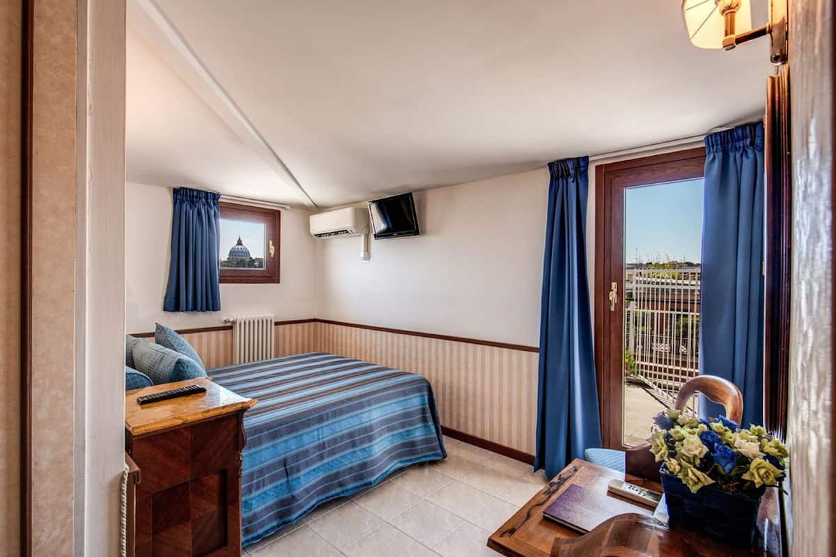 Hotellihuone, jossa on sänky, työpöytä, jossa on kaukosäädin, seinälle asennettu televisio ja ikkuna, jossa on verhot.