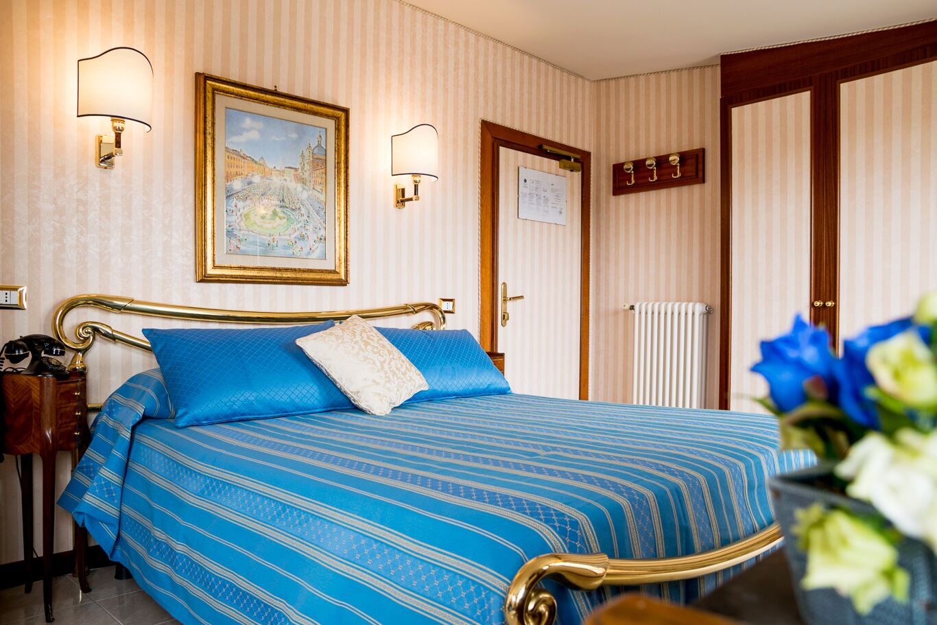A hotel room with a bed, a nightstand, a lamp, a framed picture, a door, and a radiator.