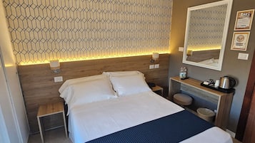 A modern hotel room with a large bed, a wooden headboard, a nightstand with a lamp, a small table, and a wall-mounted mirror.