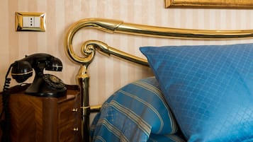 Un letto con una coperta blu e un poggiatesta dorato, un comodino in legno con un telefono rotativo nero e un interruttore della luce fissato al muro.