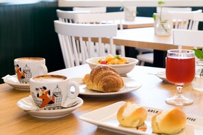 Une table à manger avec un croissant, un saladier de fruits, un verre de boisson rouge et deux tasses de café avec leurs soucoupes.