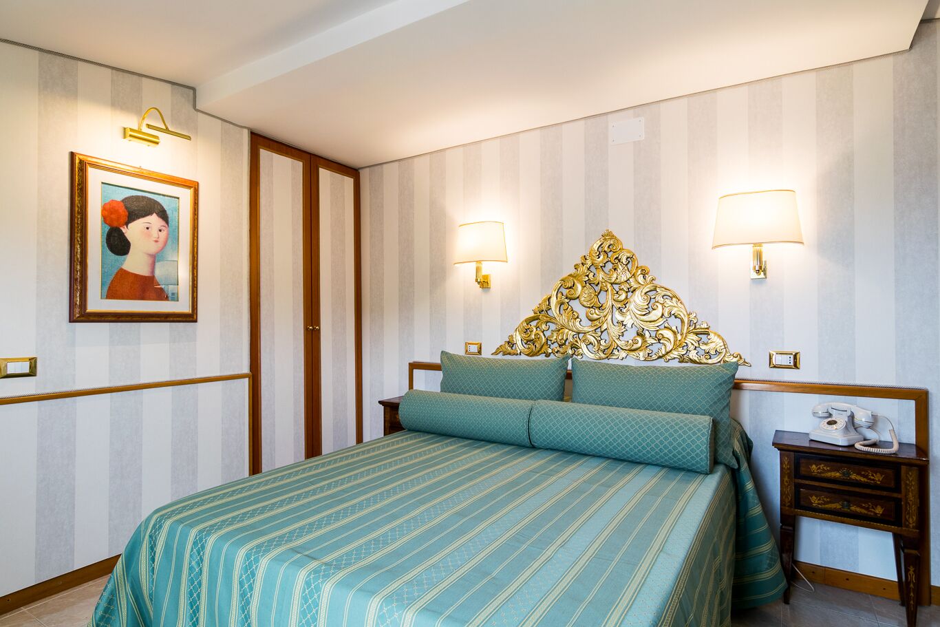 A hotel room with a bed, two bedside lamps, a nightstand with a telephone, and a framed picture on the wall.