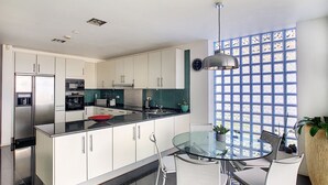 Eine moderne Küche mit weißen Schränken, einem Edelstahlkühlschrank, einer Glasinsel und einem großen Fenster mit blauem Gittermuster.