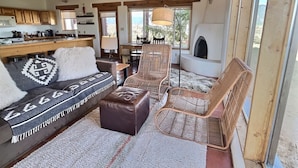A cozy living room with a sofa, two wicker chairs, a fireplace, and a kitchen area with wooden cabinets and a dining table.