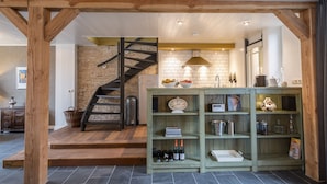 A kitchen with a brick wall, wooden beams, a staircase, and a green shelving unit with wine bottles.