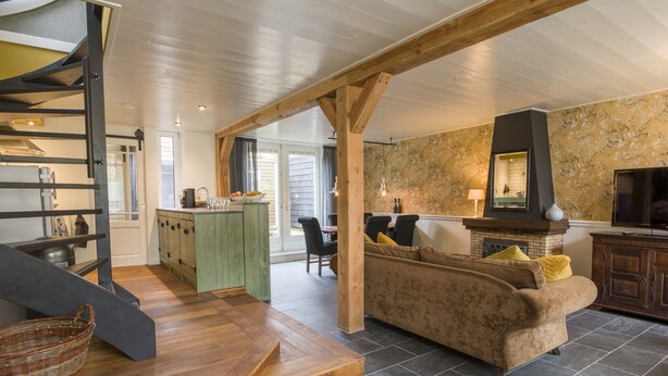 A spacious living area with a wooden staircase, a sofa, a fireplace, and a kitchen in the background.