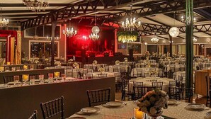 Un restaurant doté de plusieurs tables rondes dressées pour le dîner, d’une scène avec des rideaux rouges et de lustres suspendus au plafond.