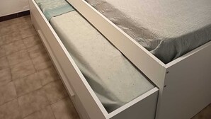 A compact bed with a storage drawer underneath, a small shelf above the bed, and a light switch on the wall.