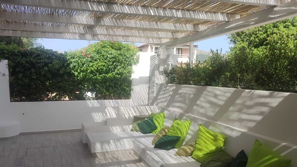 A covered outdoor seating area with a white sofa and green cushions, a tiled floor, and a view of greenery and buildings in the background.