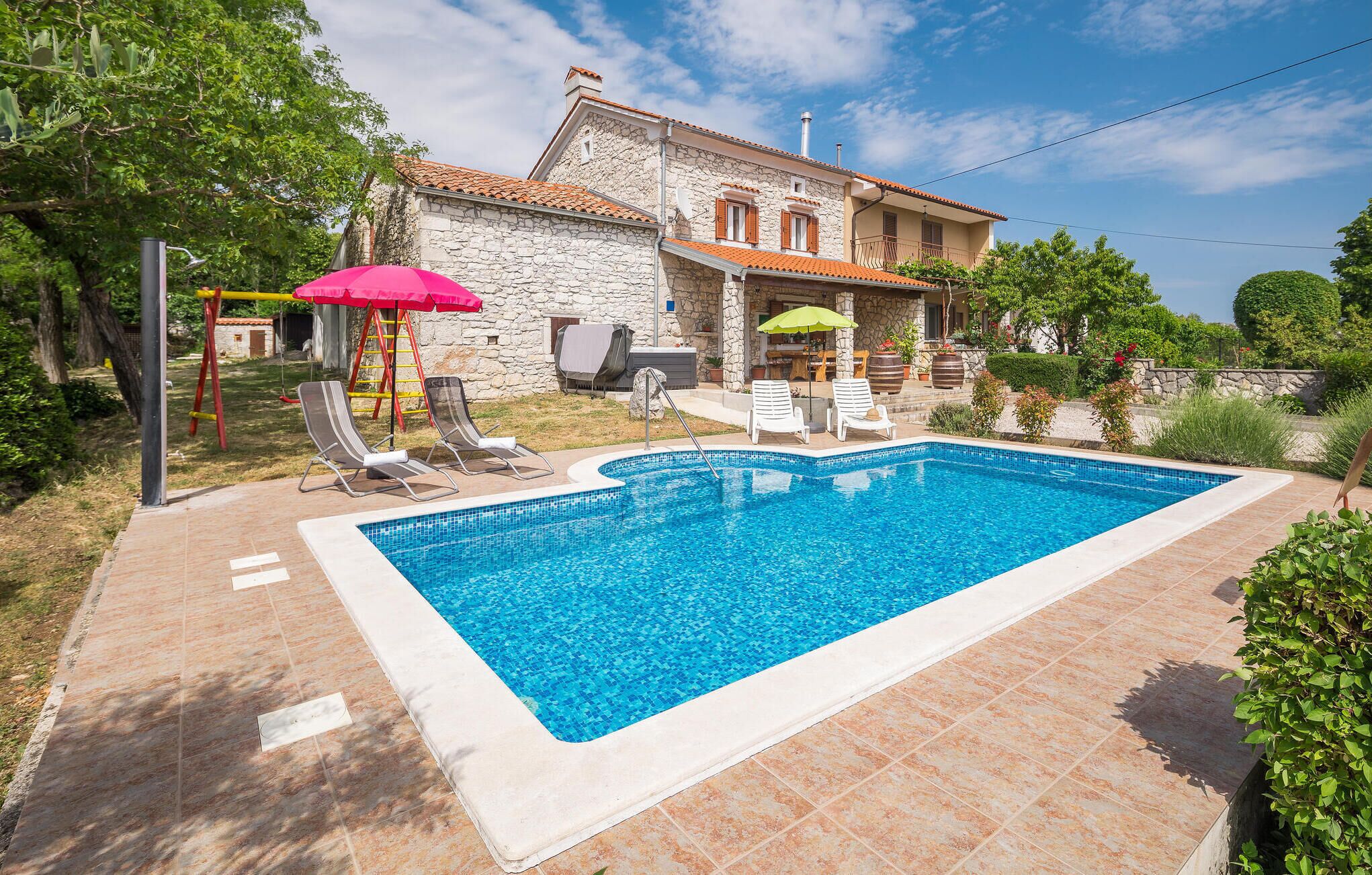 A swimming pool with lounge chairs, a stone house with a tiled roof, and a garden with trees and plants.