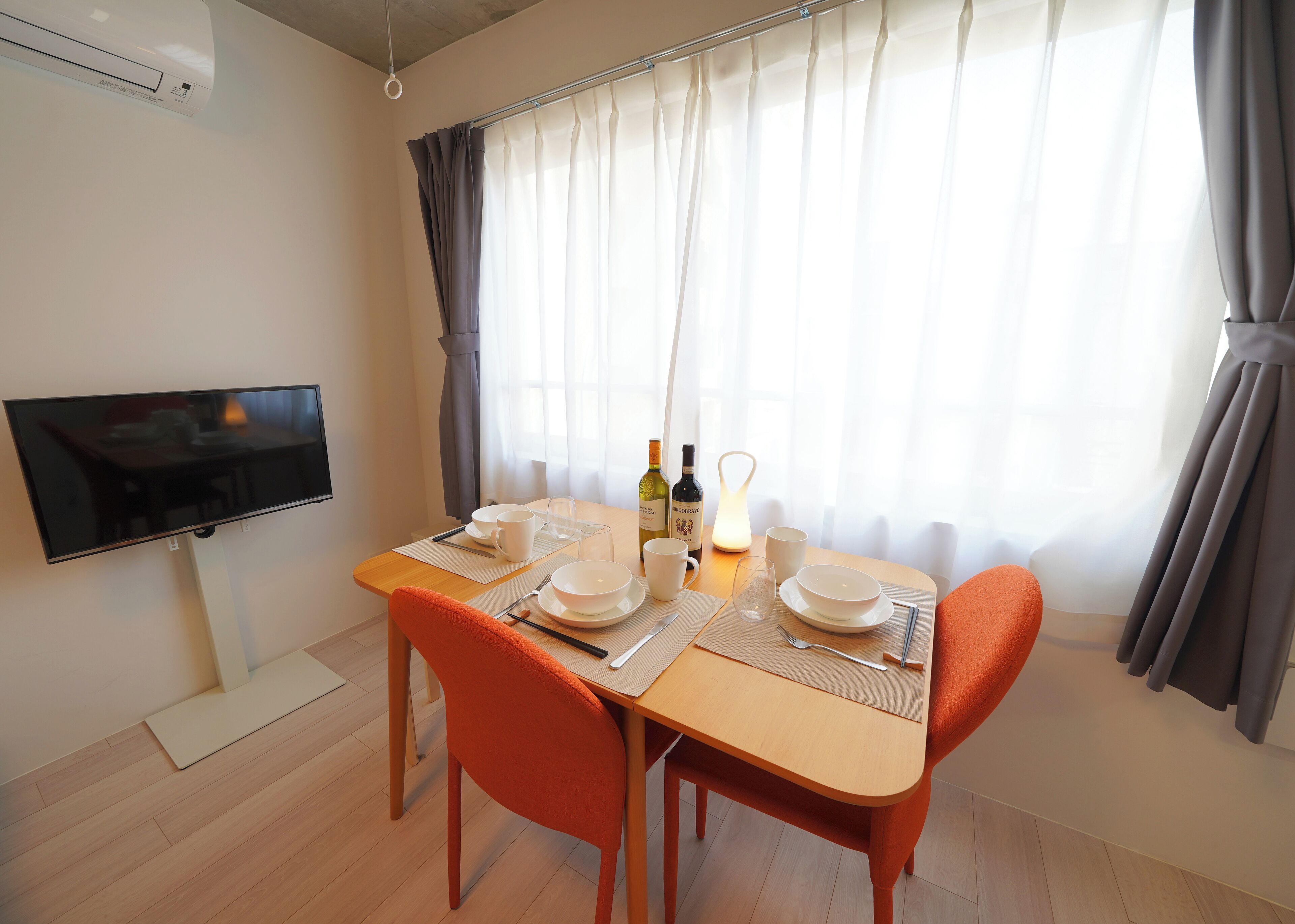 A dining area with a table set for two, a flat-screen TV mounted on the wall, and a window with sheer curtains.