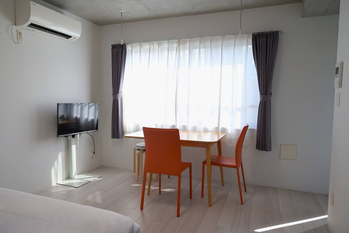 Quarto de hotel com cama, televisão, mesa de jantar com cadeiras e janela com cortinas.