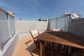 Une terrasse sur le toit, avec une table et des chaises en bois, un sol carrelé et une vue sur des immeubles et une tour en arrière-plan.