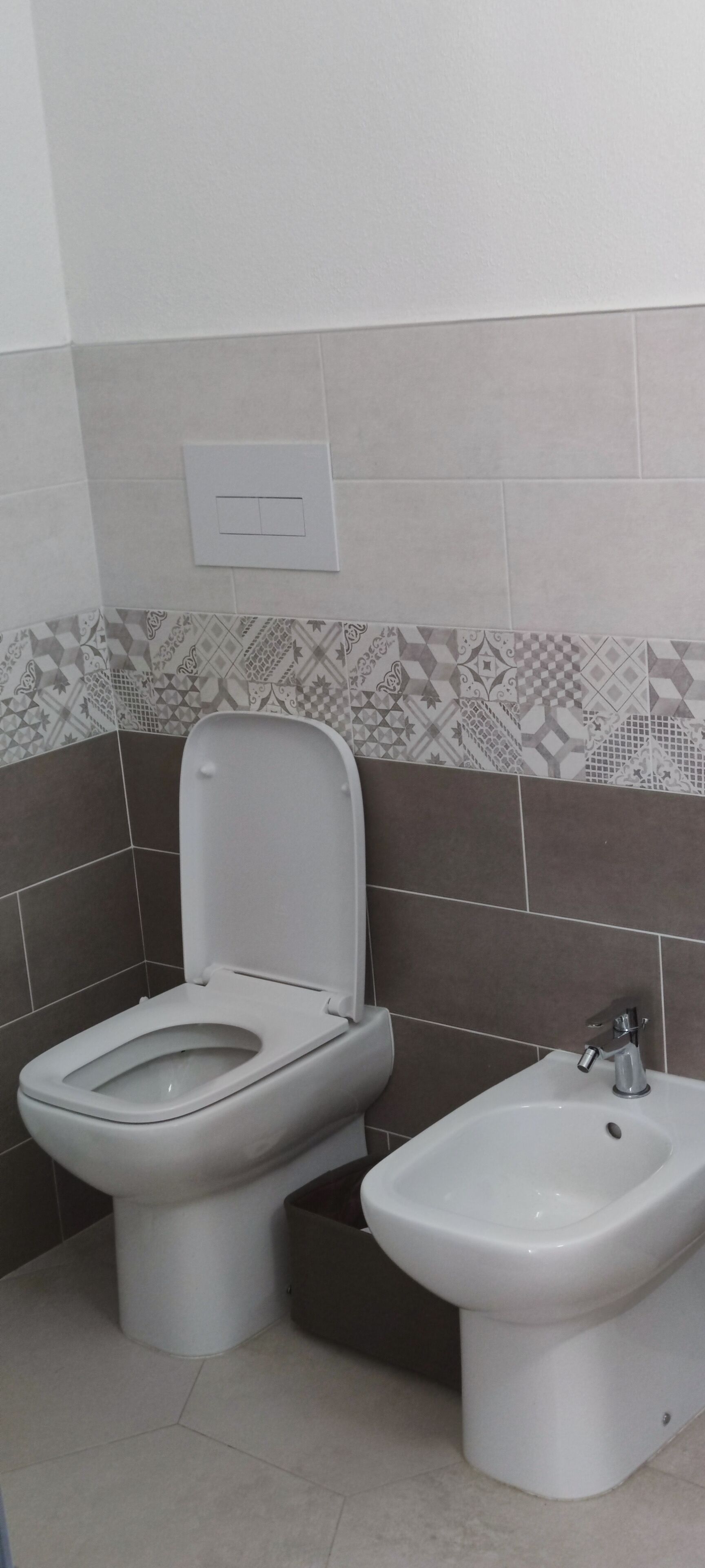 Une salle de bain avec des toilettes et un bidet, des murs carrelés et un interrupteur.