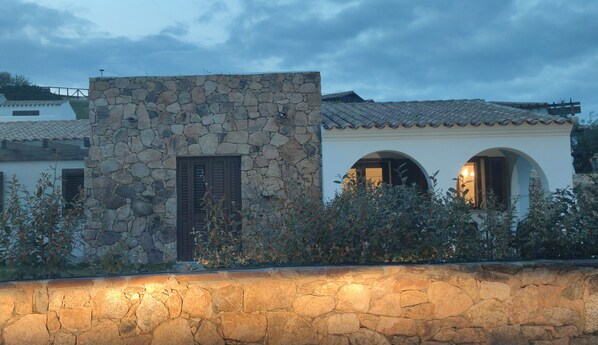 A stone building with a tiled roof, a stone wall in front, and a white building with arched windows in the background.